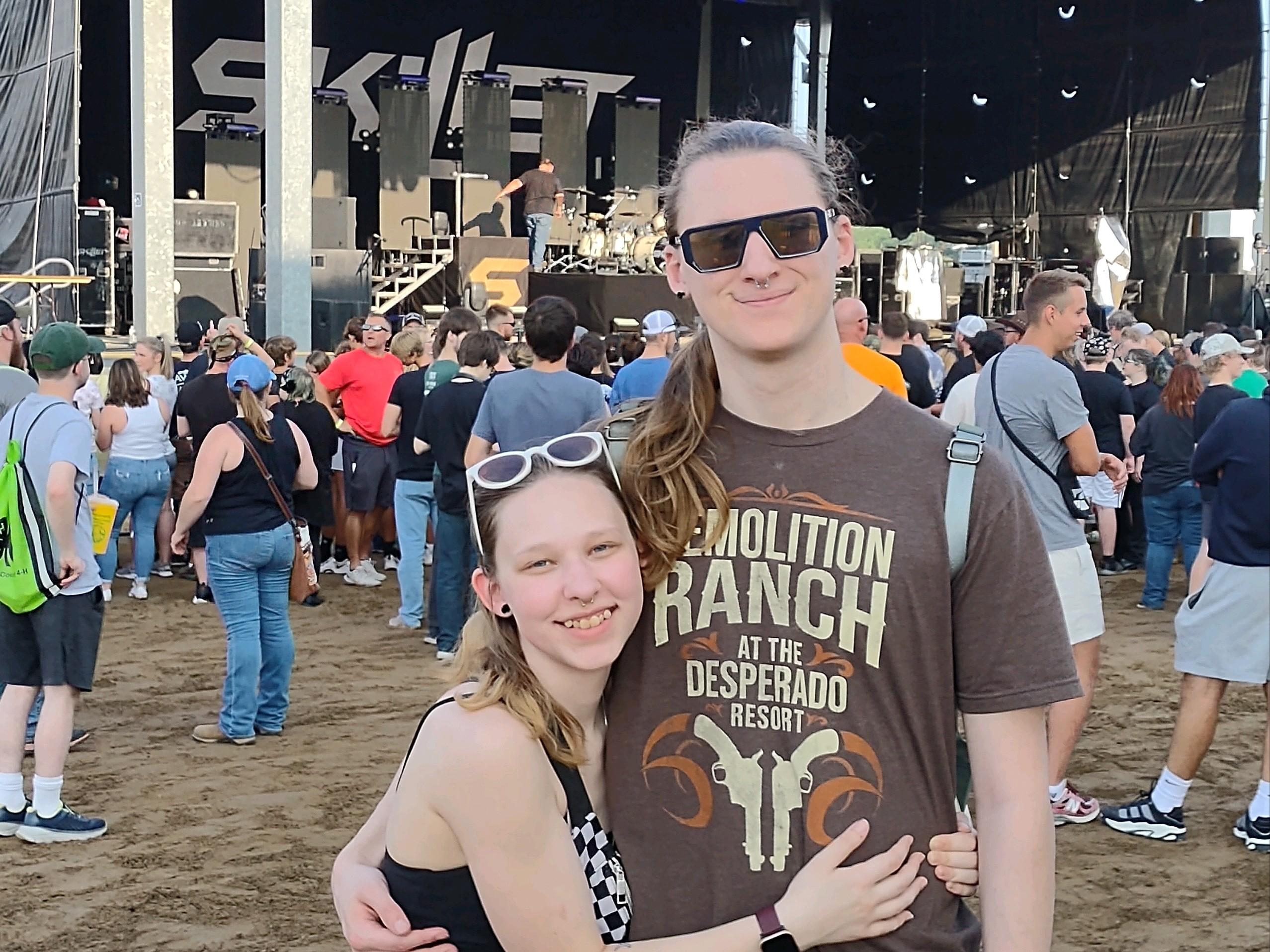 A photo of my partner (left) and I (right). We are standing side by side in front of a concert stage.